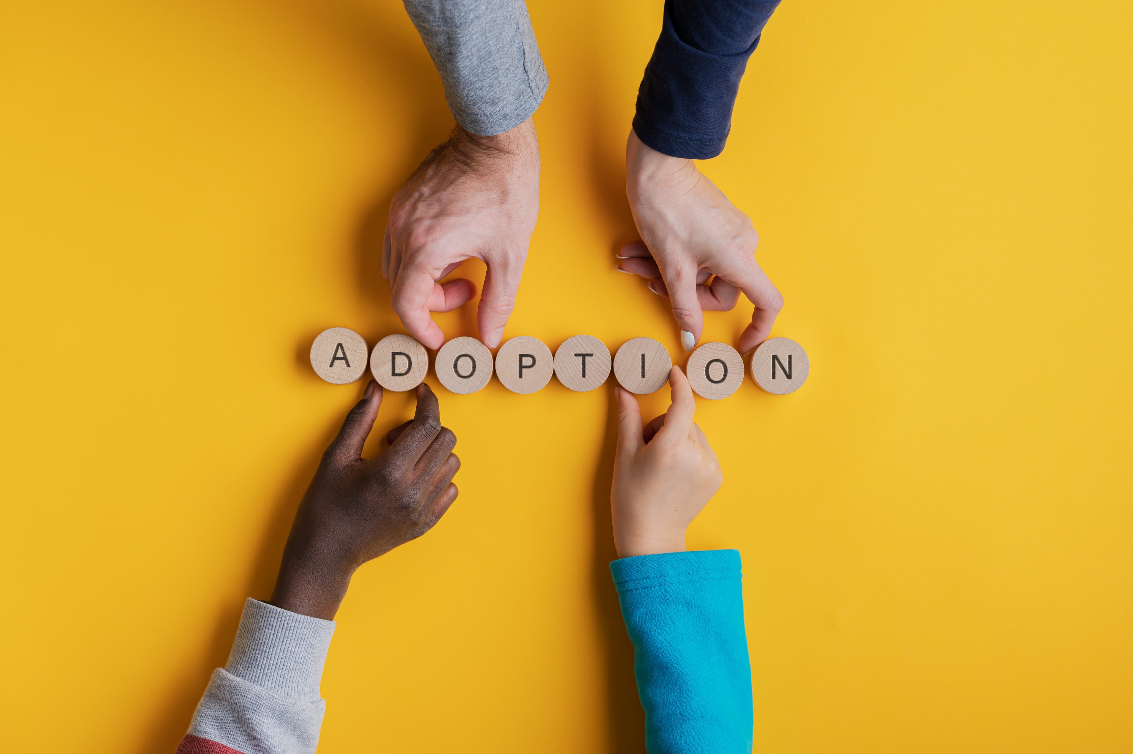 Hands of a family with black and white children assembling a word Adoption spelled on wooden cut circles. Over yellow background.