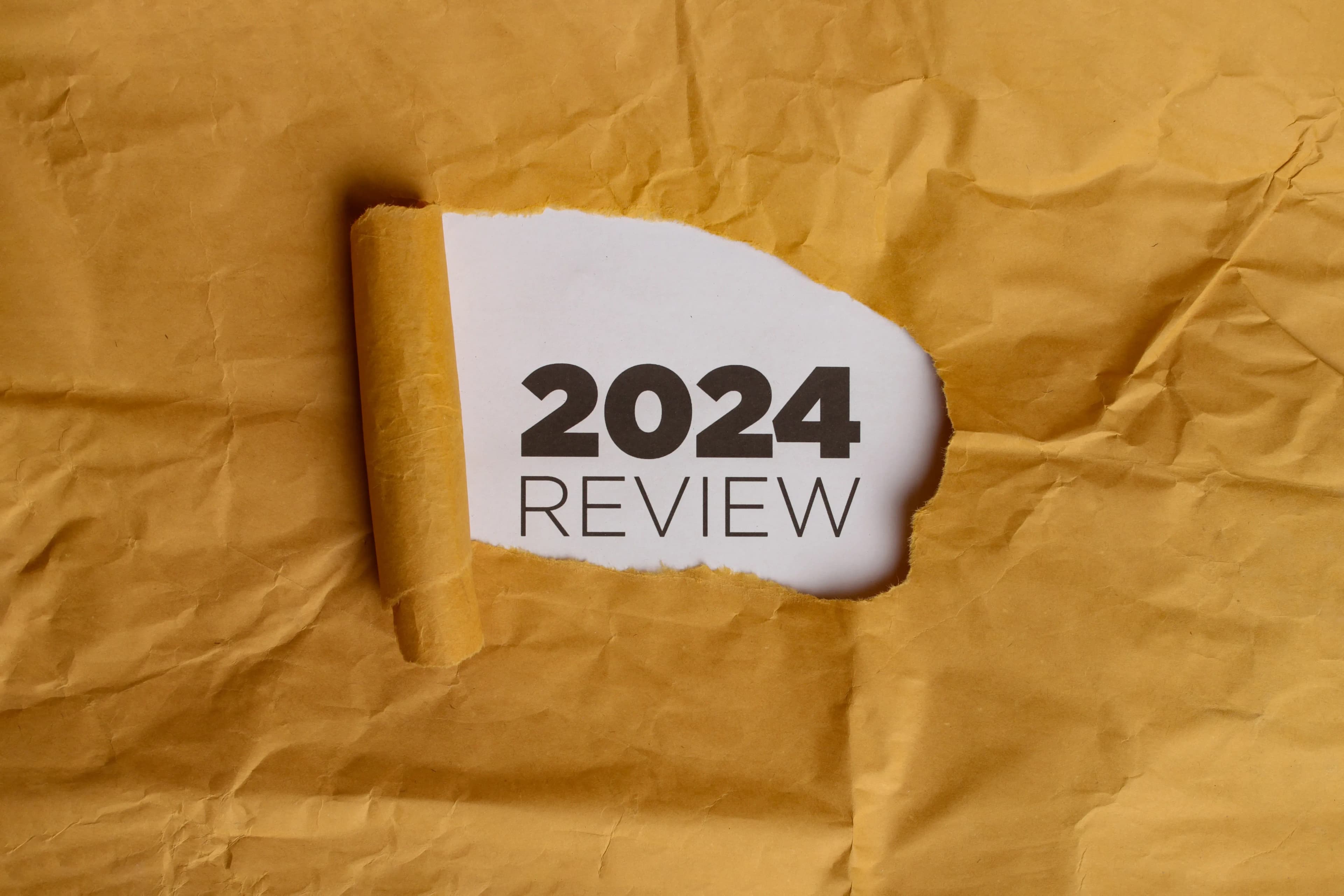 A yellow piece of paper with a hole torn in it. Through the hole, a white piece of paper is visible with the text "2024 REVIEW" written in black. This represents the concept of reviewing the year 2024