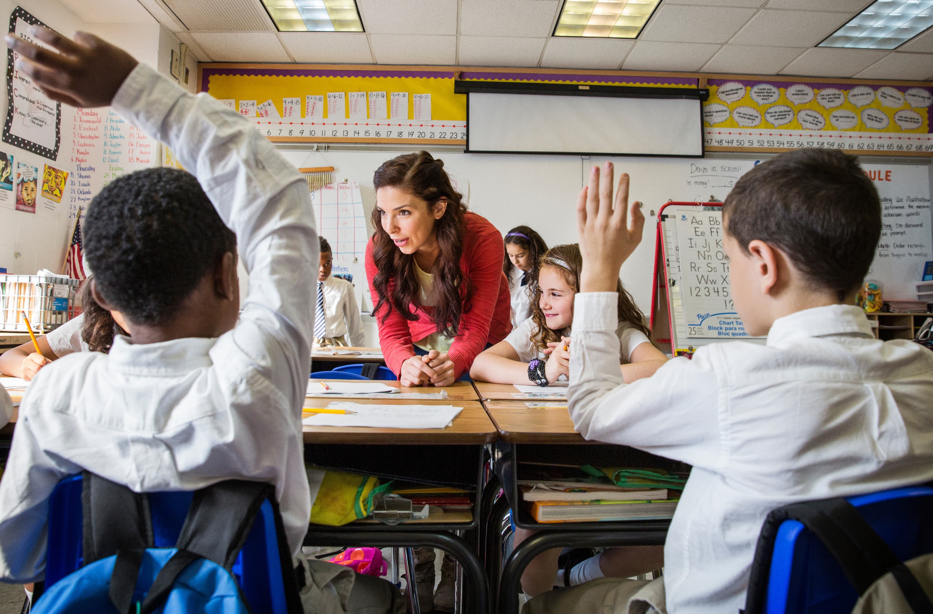 Portrait of female teacher in classroom with two schoolboys (8-9 and 10-11) raising hands in foreground