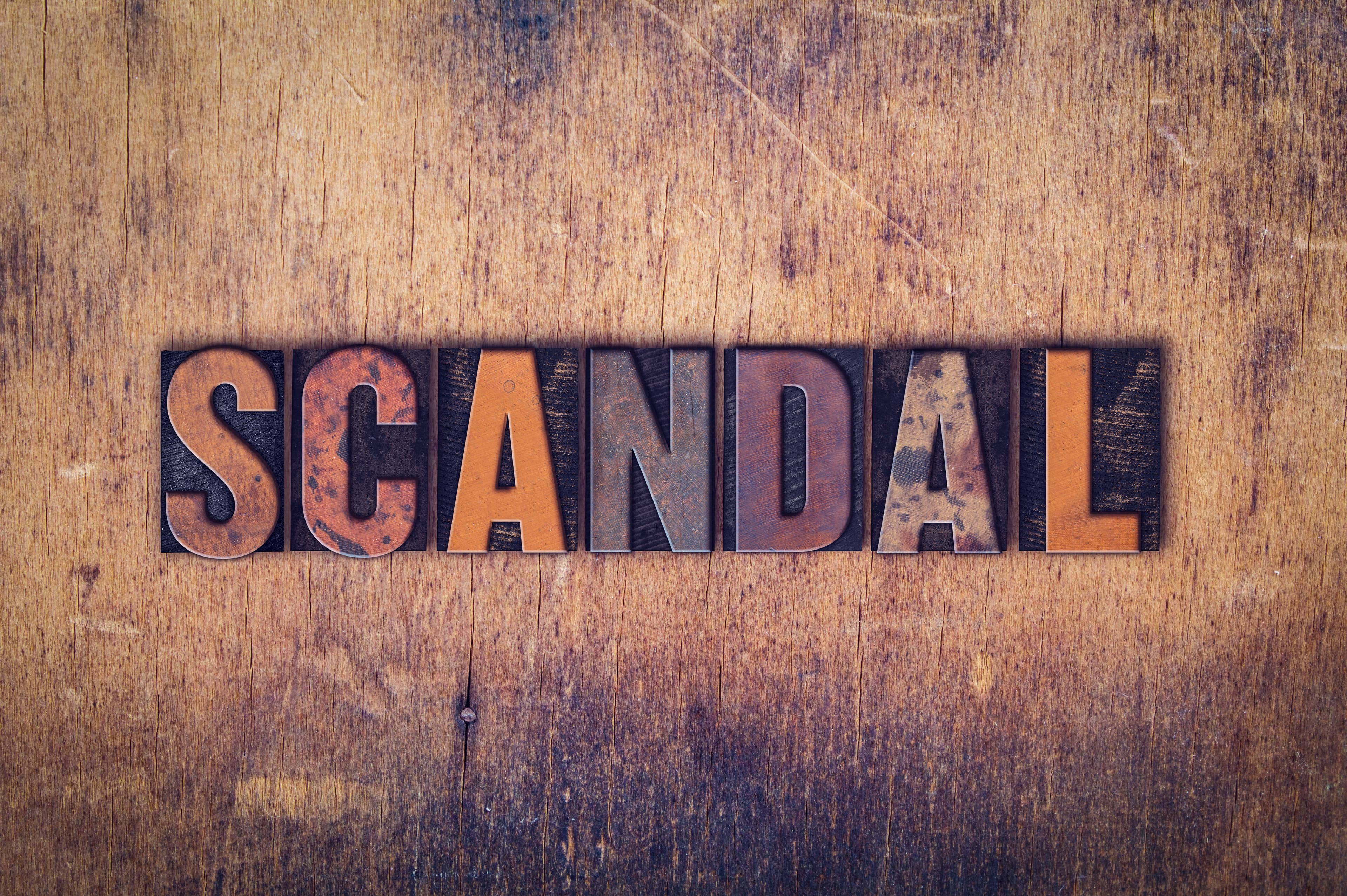 The word "Scandal" written in dirty vintage letterpress type on a aged wooden background.