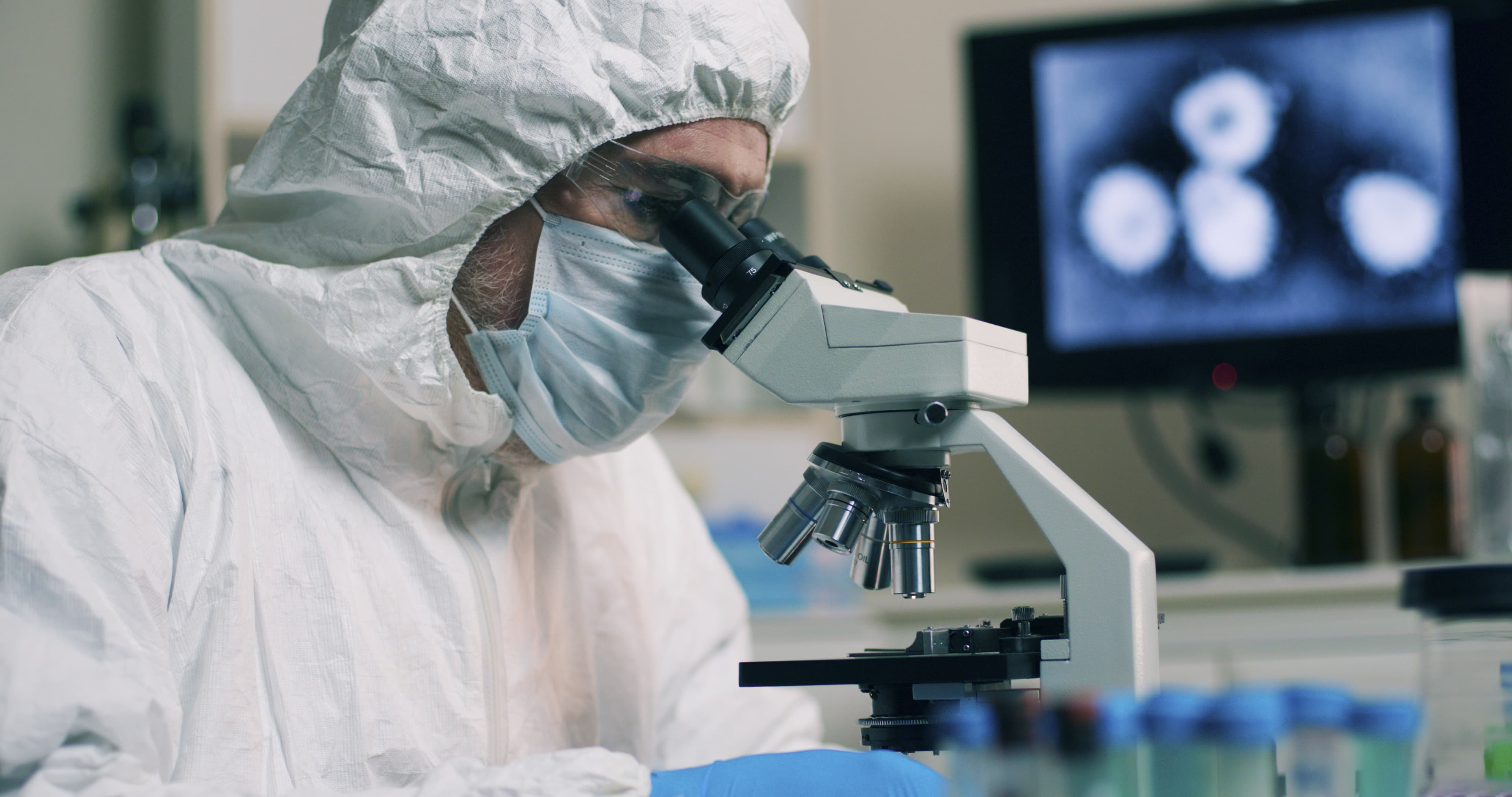 A research scientist working in his laboratory with a sample or specimen and an image of the Wuhan Coronavirus displayed on a computer screen.