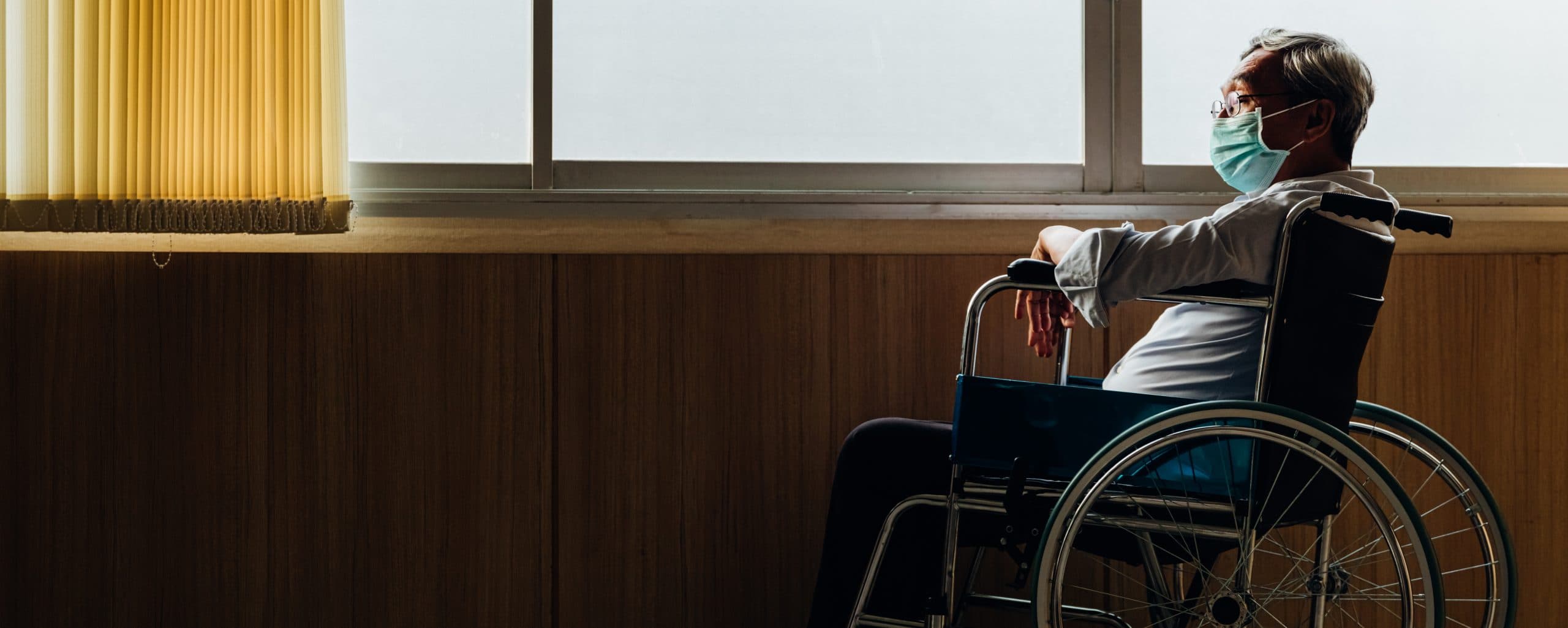 Retirement concept. Panoramic asian elderly man wear mask sitting in wheelchair while looking out the window in nursing home or hospital.