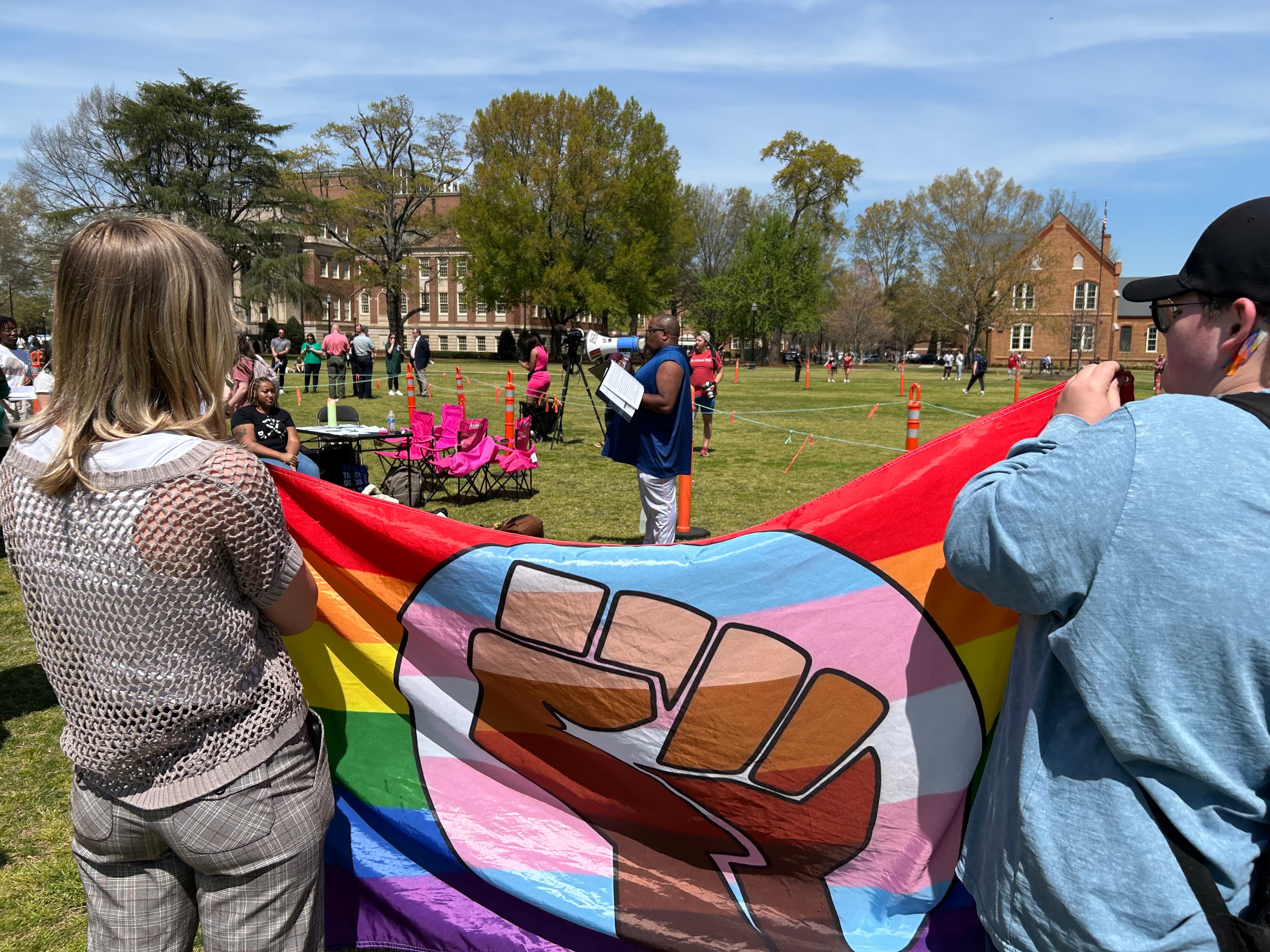Two people holding a pride flag as a speaker addresses a group of protestors using a megaphone.