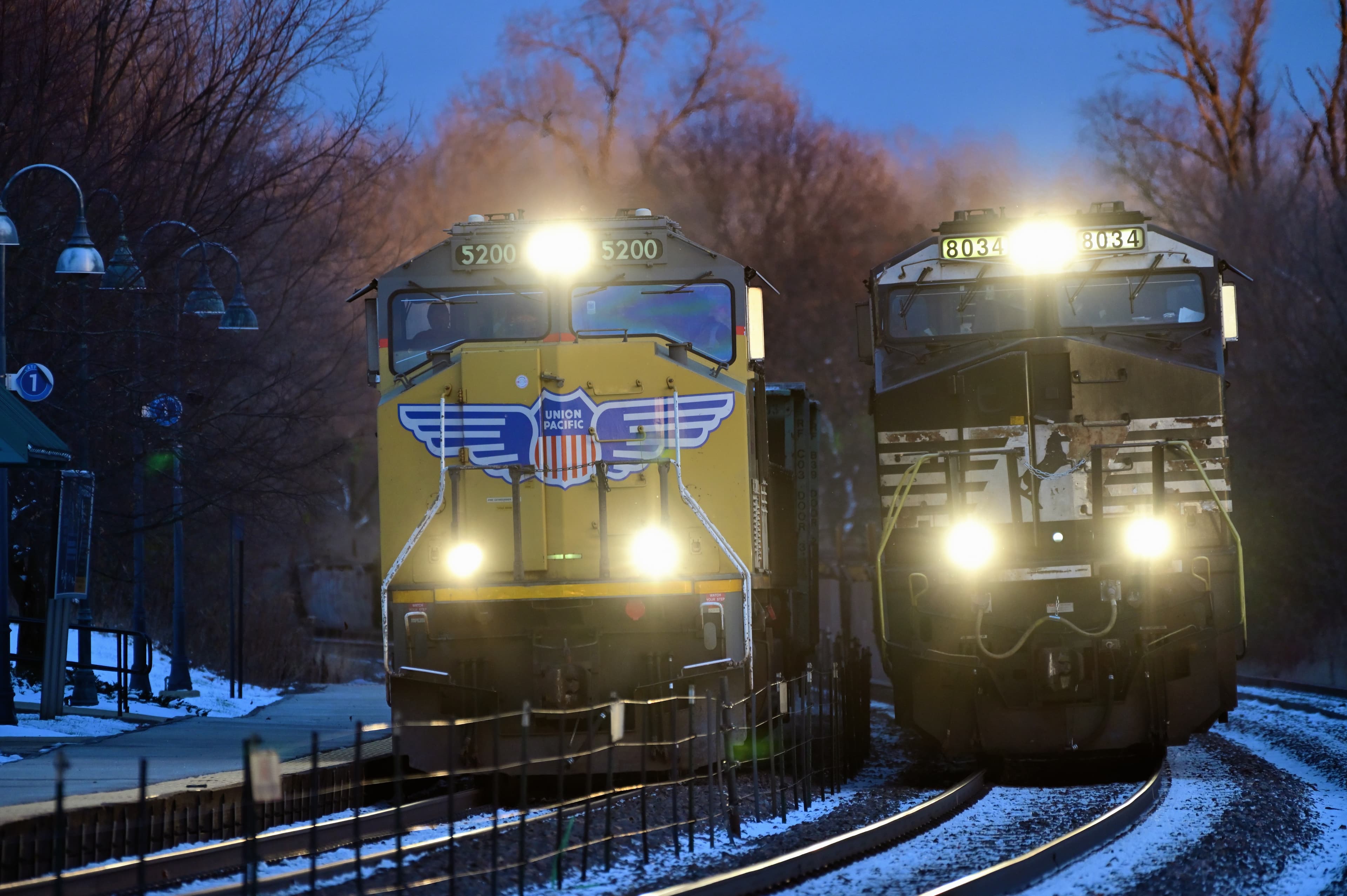 Union Pacific Railroad freight trains, one powered by an off-road Norfolk Southern Railway run-through locomotive proceed westbound, side-by-side into the the night as they pass through suburban Chicago.