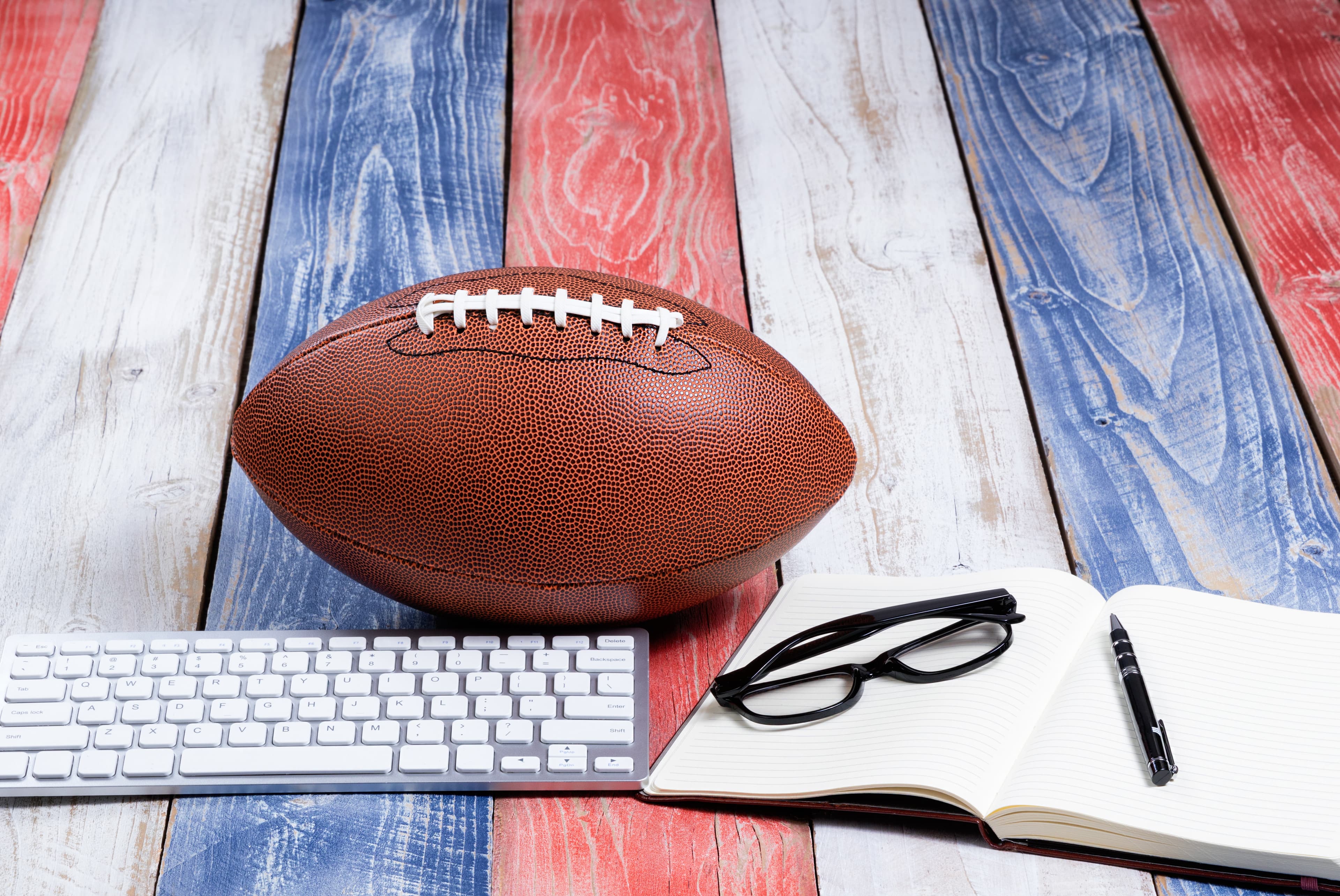 Front view of computer keyboard, reading glasses, notepad, pen and American football on red, white and blue rustic wooden boards. Concept of draft and plays for the season.
